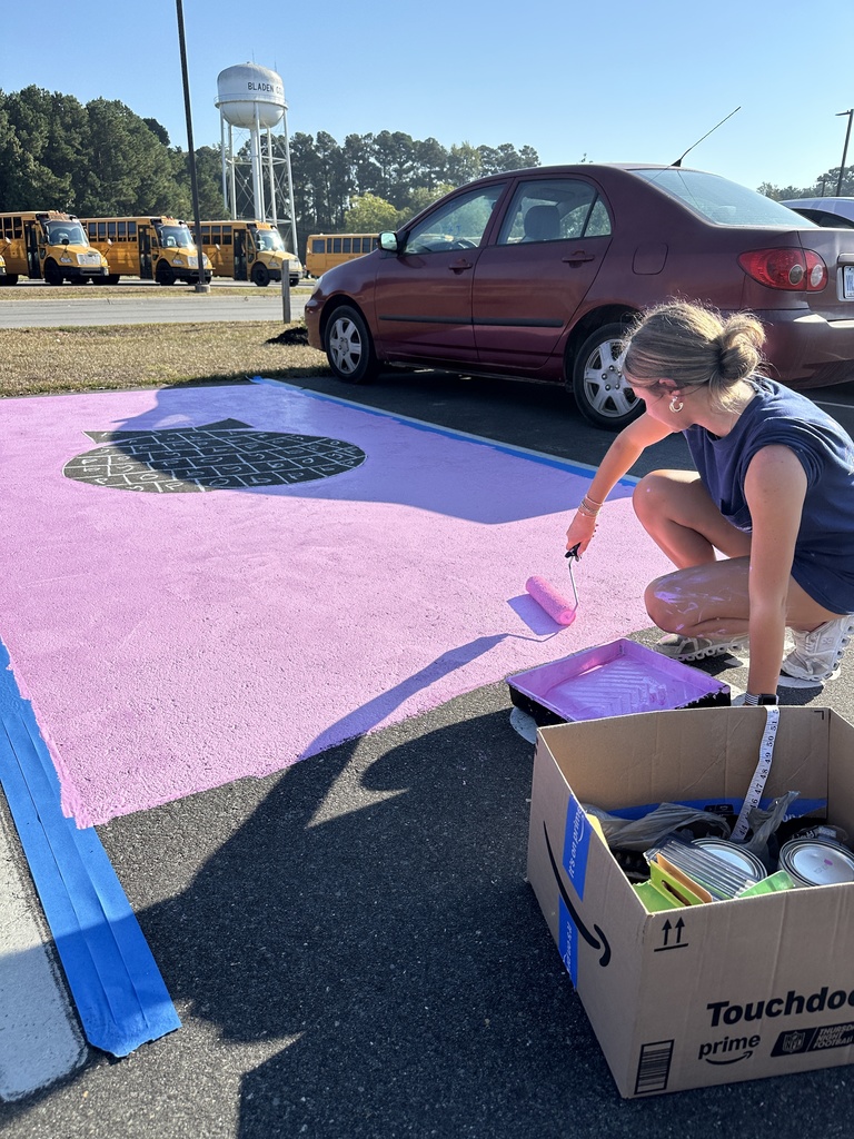 A student crouches down painting a parking spot pink with a circular black-and-white design in the center; school buses are parked in the background.