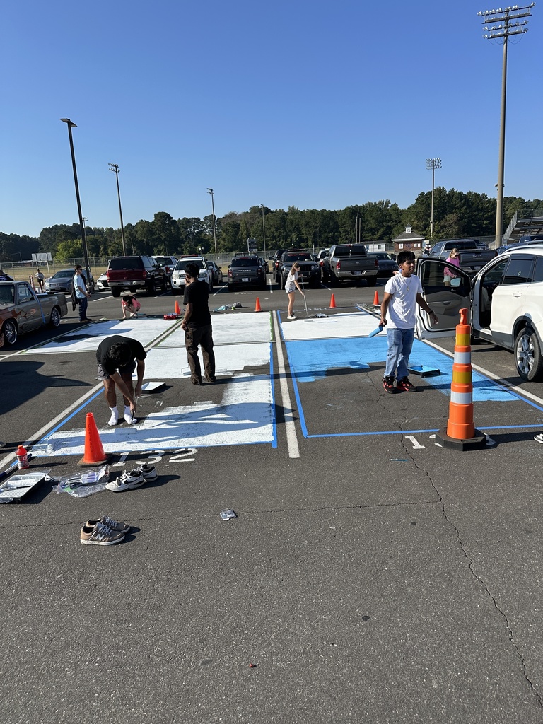 Groups of students and families work together painting large white and blue rectangles across multiple parking spaces.