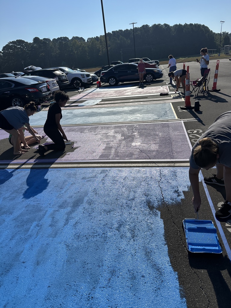 Several students use rollers to paint parking spots with blue and purple paint while others stand nearby.