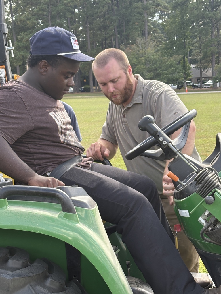 This image shows two individuals interacting with a green tractor. The person on the left, seated on the tractor, appears to be learning how to operate it, while the person on the right is standing and providing guidance or instruction. The setting looks like an outdoor field surrounded by tall trees, suggesting it could be a training or educational environment, possibly at a farm, agricultural program, or outdoor skills workshop.