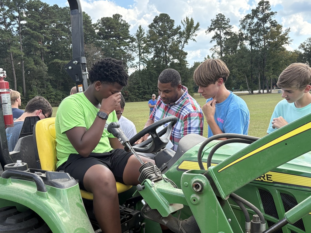 In this image, a group of students is gathered around a green John Deere tractor, receiving hands-on instruction. One student is seated in the driver’s seat, appearing focused and possibly a bit overwhelmed, while a man — likely an instructor or teacher — is providing guidance. Other students stand nearby, watching attentively and thoughtfully, indicating that this is likely an educational or training session, possibly related to agricultural studies or a vocational program.  The setting is an open grassy field with trees in the background, suggesting a rural or school farm environment. The overall mood of the image conveys engaged learning and teamwork.