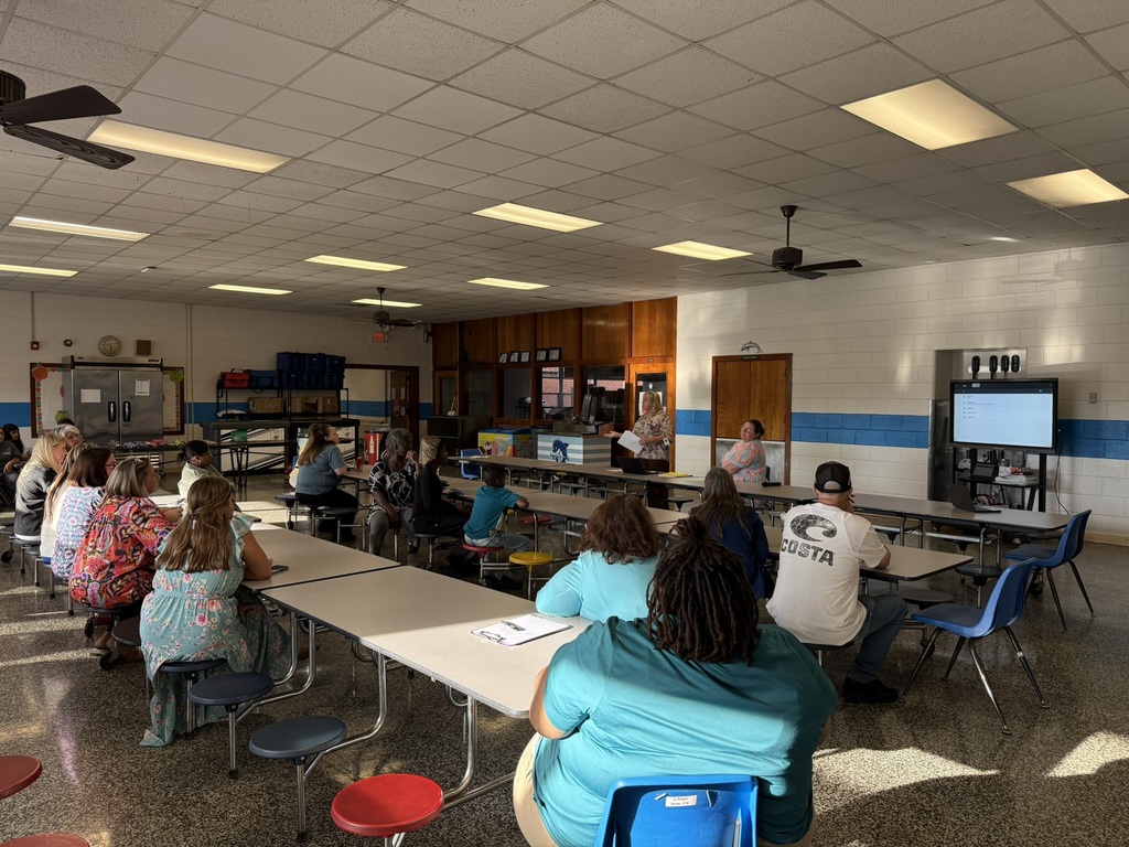 A wide view of families seated in the cafeteria tables while school staff lead the Parent Advisory Meeting.