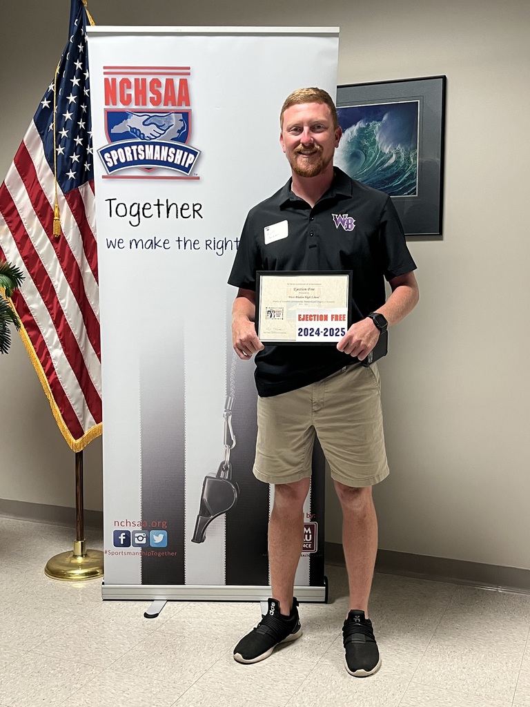 A man wearing a black polo shirt with a "WB" logo and khaki shorts stands smiling while holding a framed certificate. Behind him is a banner with the NCHSAA Sportsmanship logo and the words "Together we make the right call," along with an American flag. The certificate reads “Ejection Free 2024–2025.”