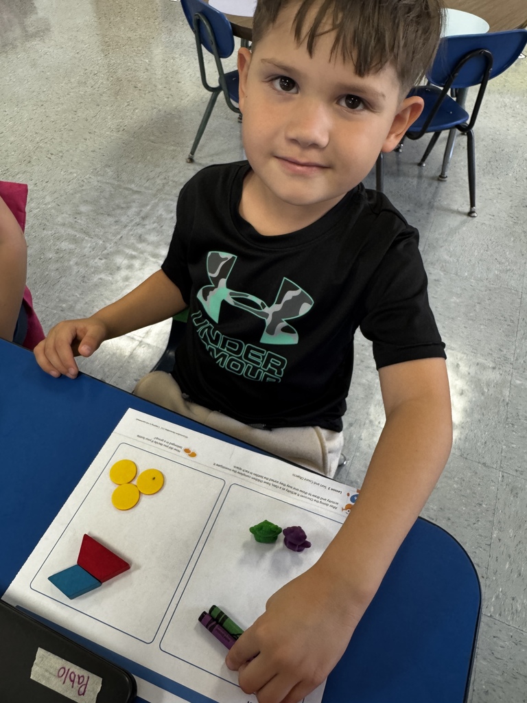 Student in a black Under Armour shirt sits with a worksheet showing red, green, and purple counters and crayons.