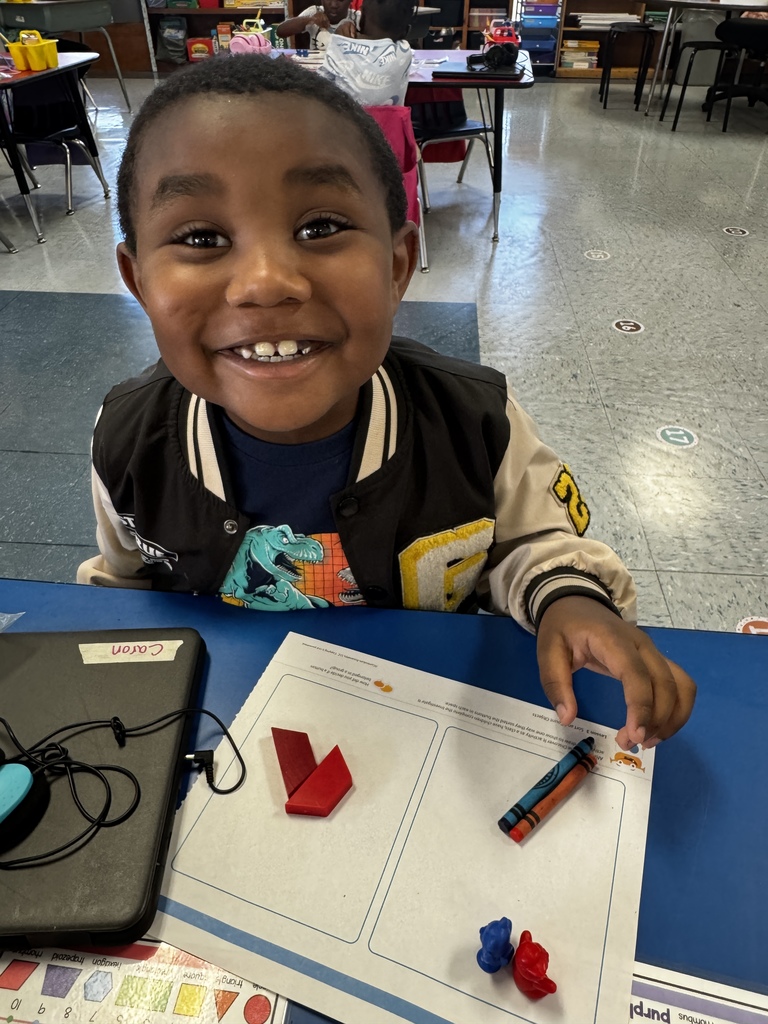 Child in a black and white jacket grins while pointing to red and blue math manipulatives on a worksheet.