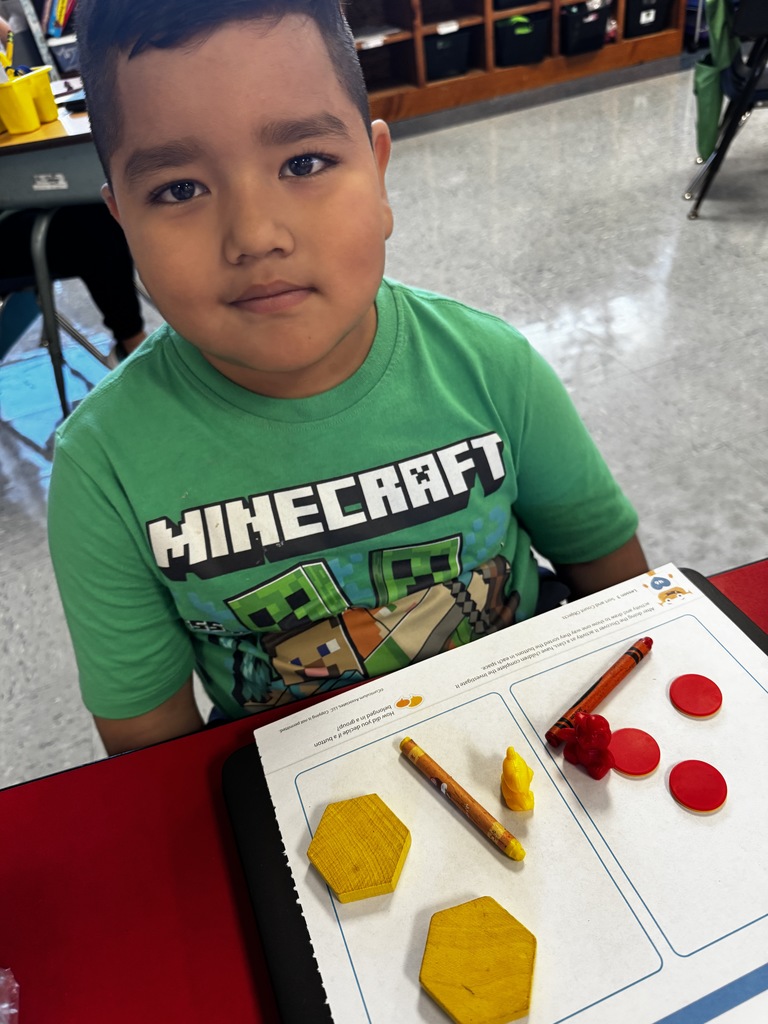 Student in a green Minecraft shirt sits at a desk with yellow blocks, red counters, and orange crayons on a worksheet.