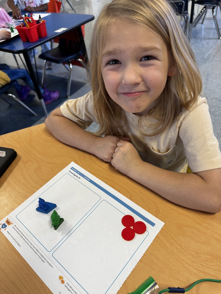 Student with long blond hair smiles while showing red and green math manipulatives on a worksheet.