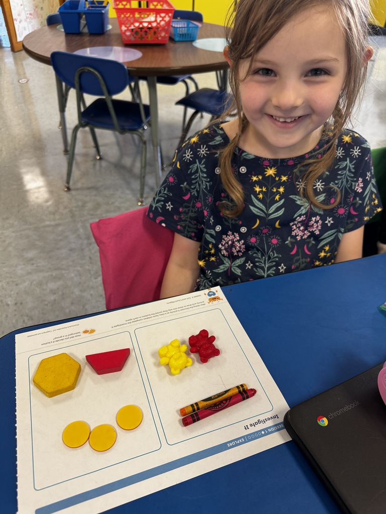 Smiling student in a floral shirt displays a worksheet with yellow shapes, red counters, and bear counters.