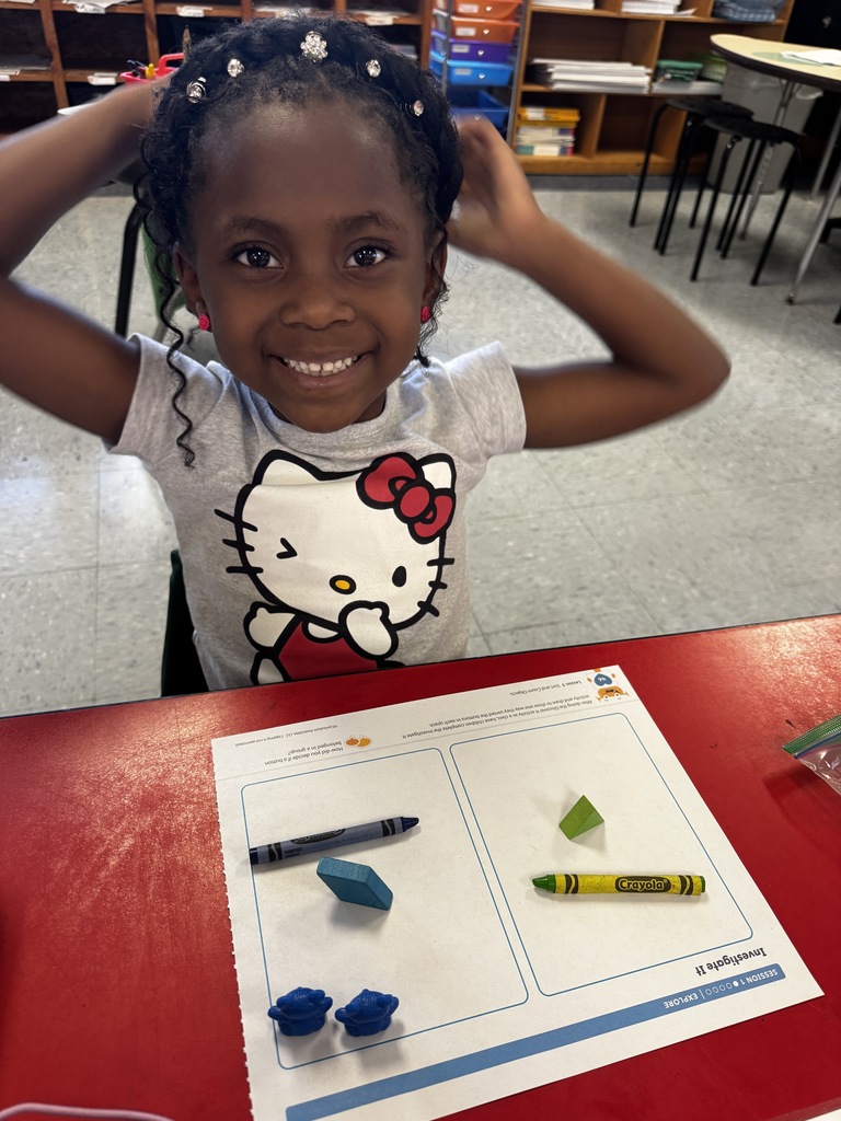 Smiling student in a Hello Kitty shirt shows a worksheet with blue and green blocks and crayons.