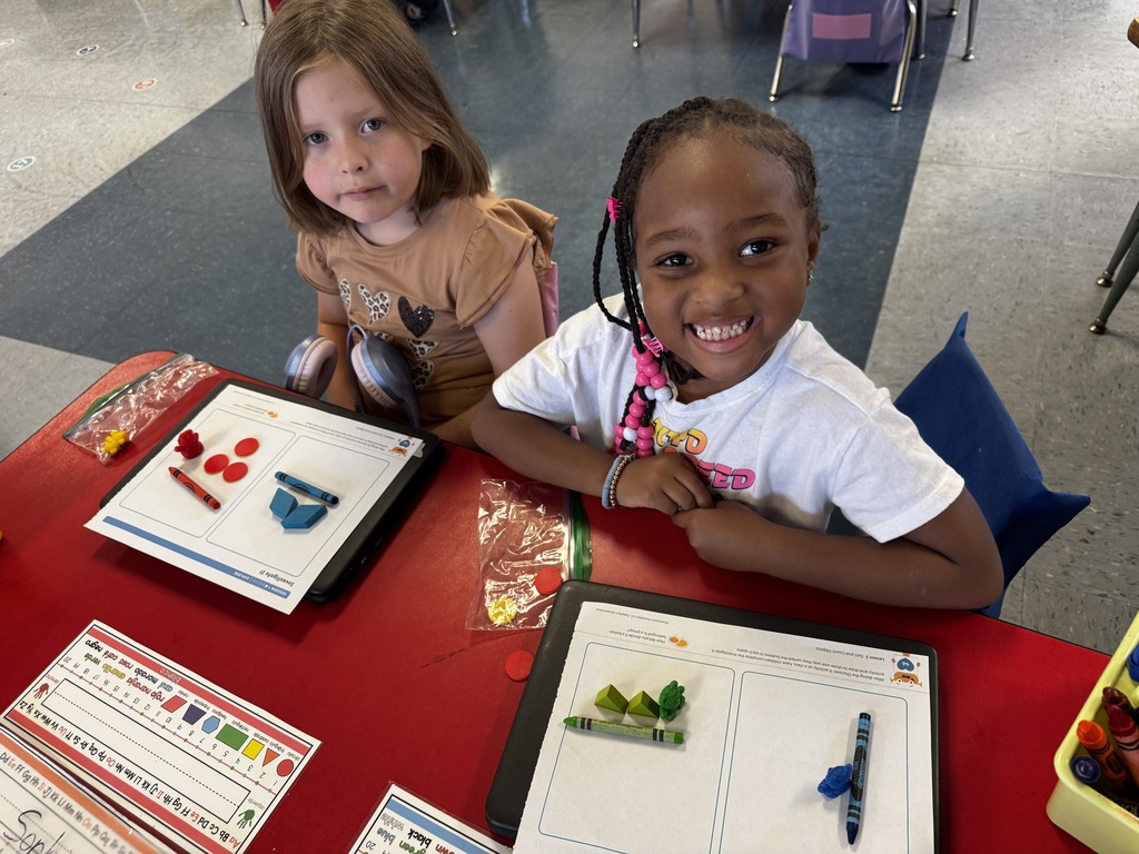 Two students sit side by side, smiling, with math worksheets covered in red and blue counters and crayons.