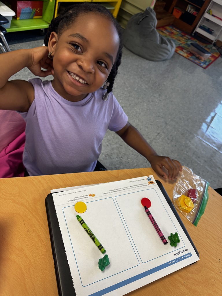 Smiling student in a purple shirt shows a worksheet with colored counters, crayons, and small math manipulatives.