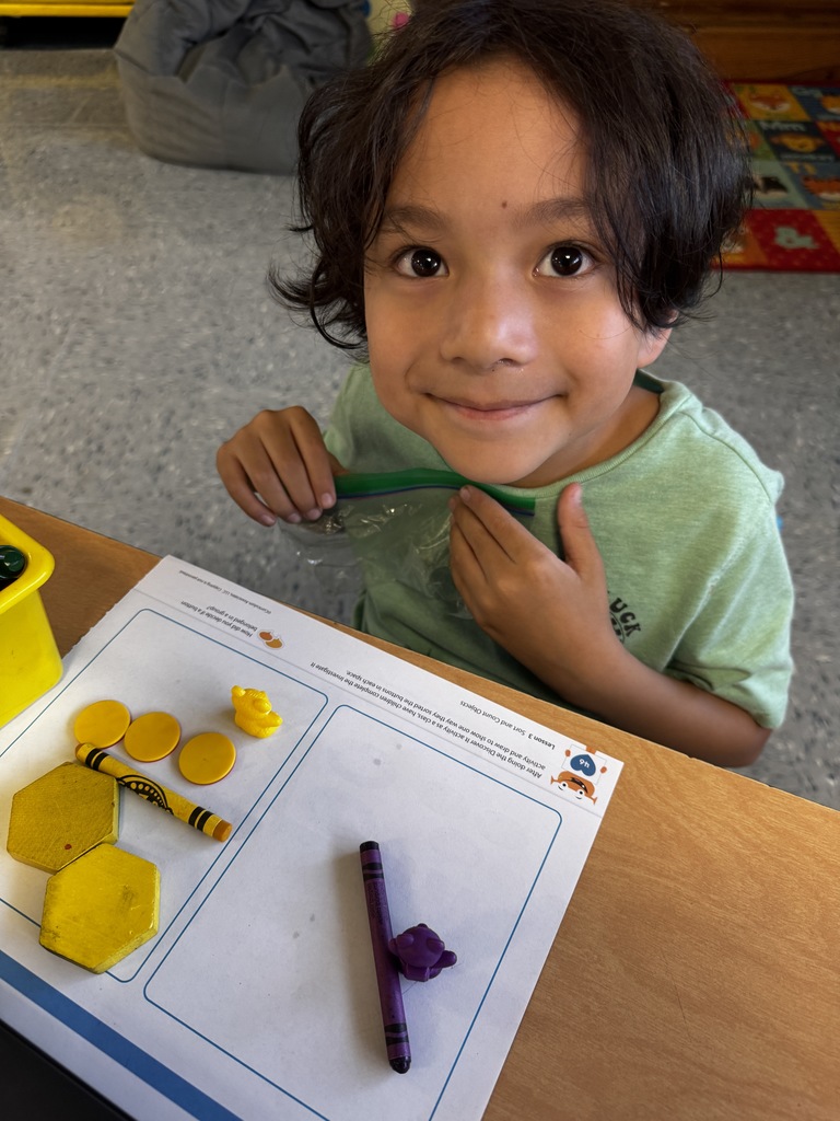 Child in a green shirt looks up with a smile while working with yellow counters and a purple crayon on a math worksheet.