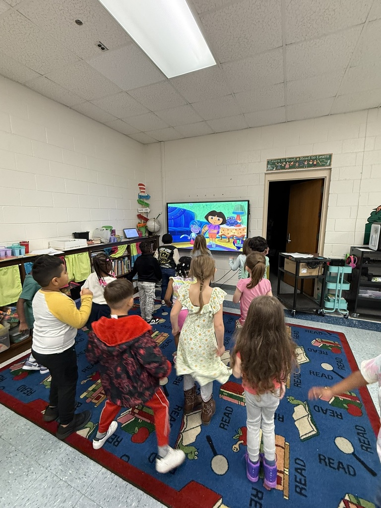 Students stand on a colorful classroom rug, moving and dancing along with a cartoon video playing on a large screen at the front of the room.