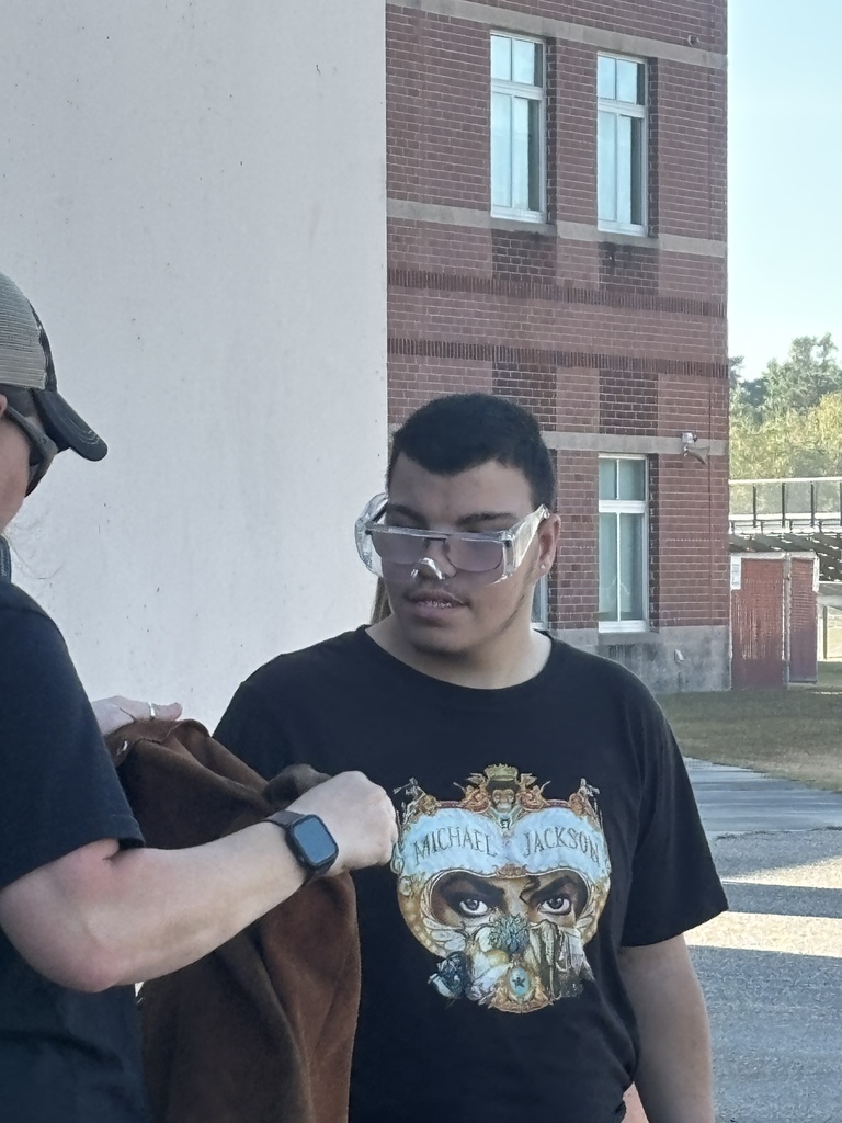 Several students in safety goggles and work clothes using a red fire extinguisher to put out a small controlled fire outdoors. An instructor stands nearby supervising while smoke rises as the flames are extinguished