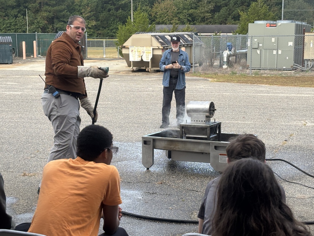 Several students in safety goggles and work clothes using a red fire extinguisher to put out a small controlled fire outdoors. An instructor stands nearby supervising while smoke rises as the flames are extinguished