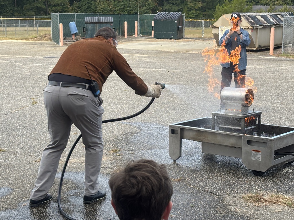 Several students in safety goggles and work clothes using a red fire extinguisher to put out a small controlled fire outdoors. An instructor stands nearby supervising while smoke rises as the flames are extinguished