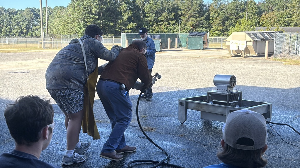 Several students in safety goggles and work clothes using a red fire extinguisher to put out a small controlled fire outdoors. An instructor stands nearby supervising while smoke rises as the flames are extinguished