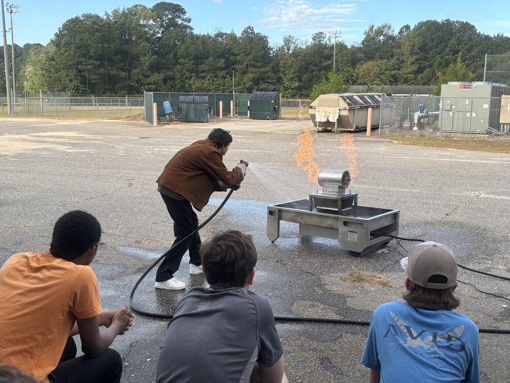 Several students in safety goggles and work clothes using a red fire extinguisher to put out a small controlled fire outdoors. An instructor stands nearby supervising while smoke rises as the flames are extinguished