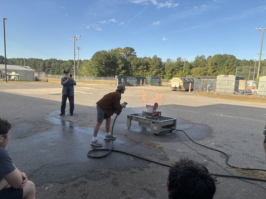 Several students in safety goggles and work clothes using a red fire extinguisher to put out a small controlled fire outdoors. An instructor stands nearby supervising while smoke rises as the flames are extinguished