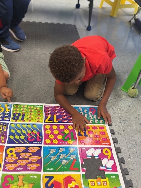 A child in a red shirt leans over a colorful floor mat with numbers and pictures, pointing to a section.