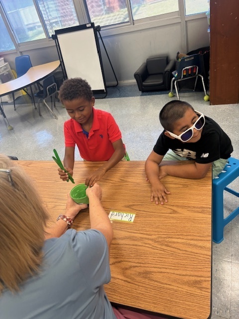 Two students sit at a table with a teacher, using large green counting sticks for a math task.