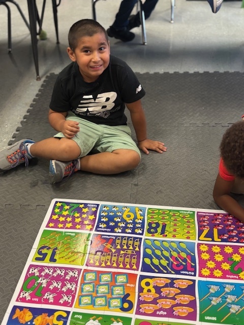 Smiling student sits beside the number mat during math playtime.