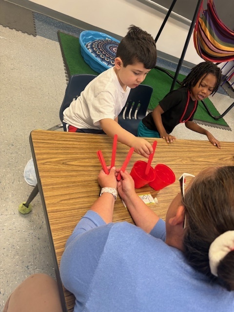 A student and teacher work together using red counting sticks during a hands-on math activity, while another student looks on.