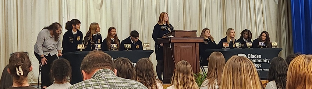 Students in FFA jackets sit at two tables on a stage during a ceremony at Bladen Community College. A student stands at the podium in the center speaking, while others are seated behind tables with FFA emblems displayed. Audience members are visible in the foreground, watching the presentation