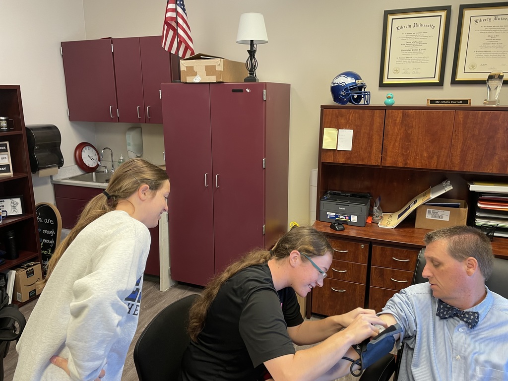 This image shows a scene inside an office where a woman is taking a man's blood pressure using a digital blood pressure monitor. Another girl stands nearby, observing the process with interest. The man is seated and dressed in a blue shirt with a polka-dot bow tie, and the woman taking the reading is wearing glasses and a black shirt.  In the background, we can see:  A cabinet and sink area, suggesting this might be a school nurse's or administrative office.  Two diplomas from Liberty University on the wall.  An American flag on top of the cabinet.  A blue football helmet and a rubber duck on the wooden shelving unit.  A sign on the desk that says "Dr. Chris Carroll," indicating this may be his office.  The atmosphere appears educational and casual, likely part of a learning activity or health awareness event.