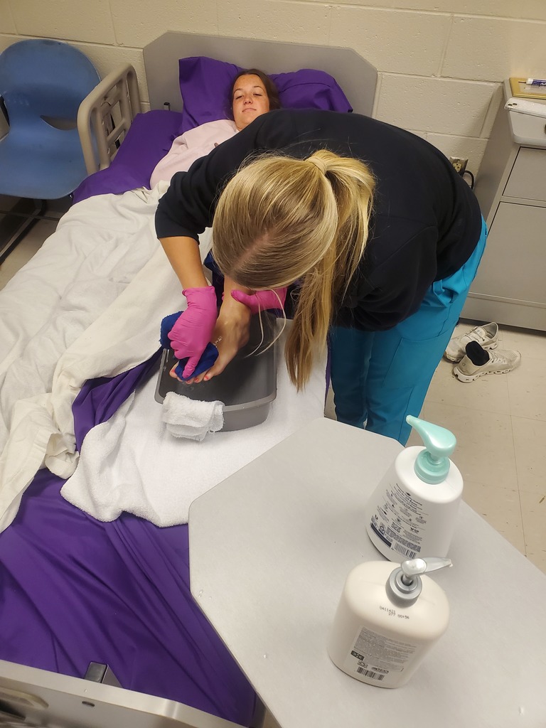 The image shows a healthcare setting where a caregiver is providing foot care to a patient lying in bed. The caregiver is wearing gloves (pink in color) and is focused on washing or cleaning the patient’s feet using a basin. The patient looks relaxed and is lying on a hospital-style bed with purple sheets and white towels for cleanliness and comfort. Nearby, on a bedside table, there are pump bottles—likely soap and lotion—used for hygiene and skin care. This appears to be a training or clinical environment, possibly a nursing or caregiver skills lab, given the setup and attention to hygiene and proper technique.
