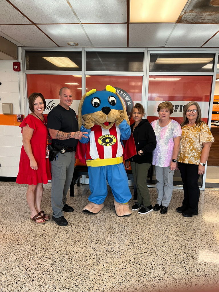A small group of adults, including a deputy sheriff, poses with the Safety Pup mascot inside a school lobby.
