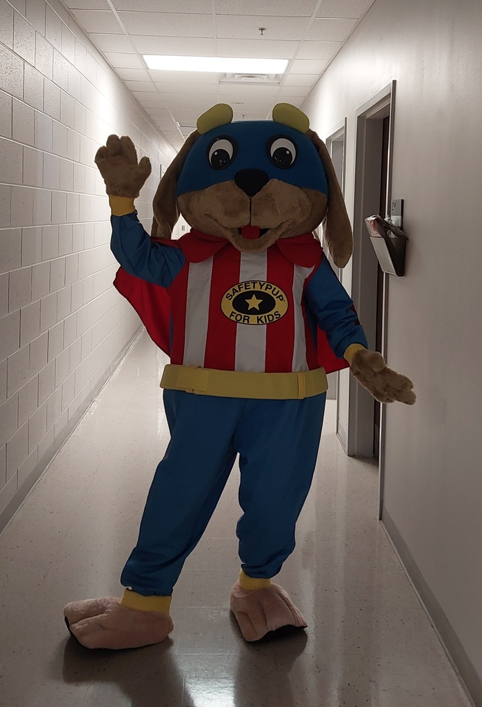 The Safety Pup mascot waves while standing in a brightly lit school hallway.