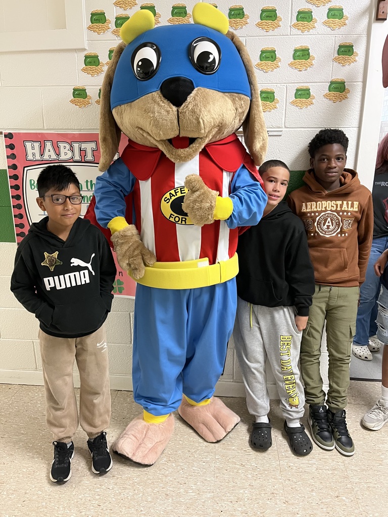 Four male students stand smiling beside the Safety Pup mascot in a school hallway decorated with student work.