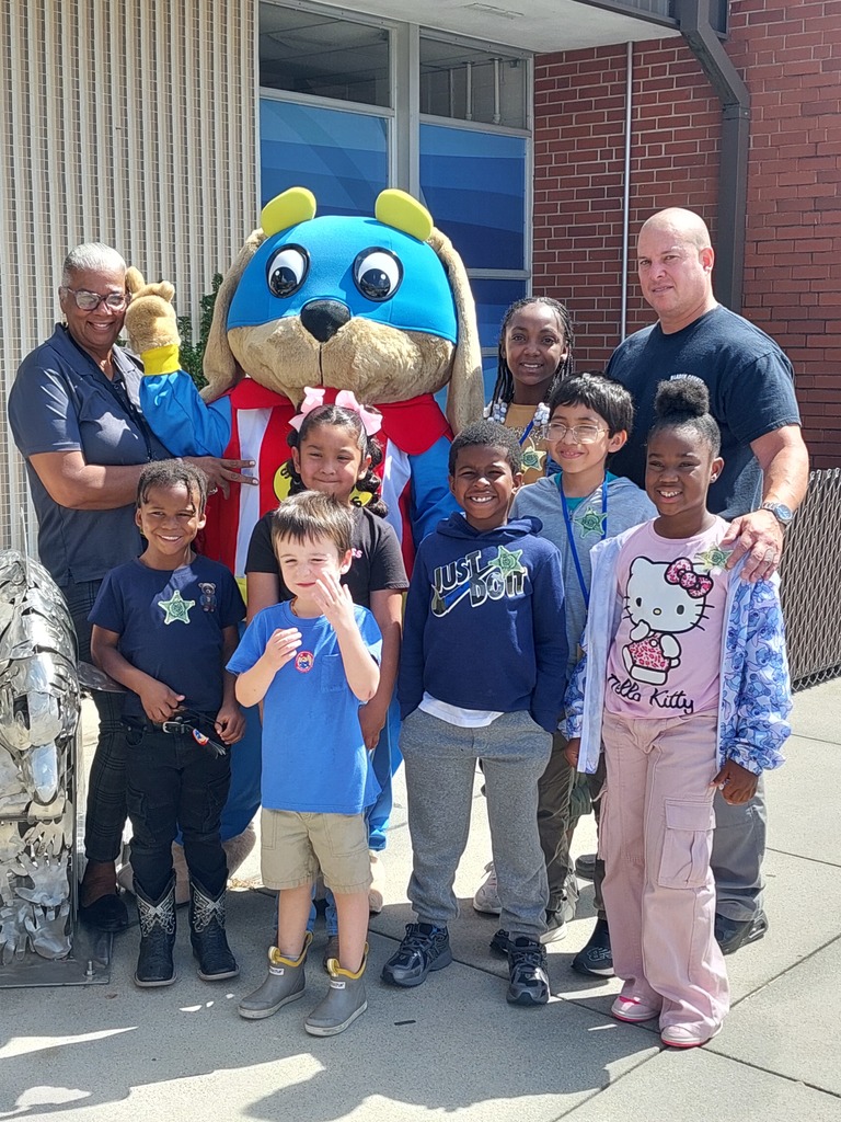 A group of elementary students and staff smile outside a school building, standing with the Safety Pup mascot.