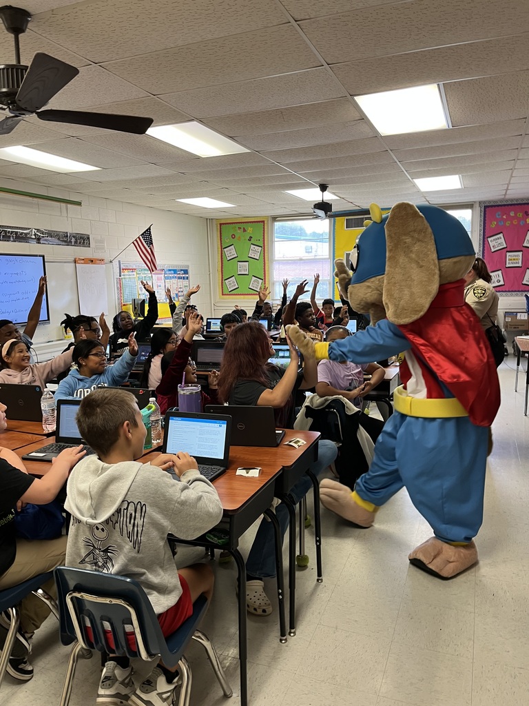 The Safety Pup mascot interacts with students in a classroom. Many students at desks raise their hands while working on laptops