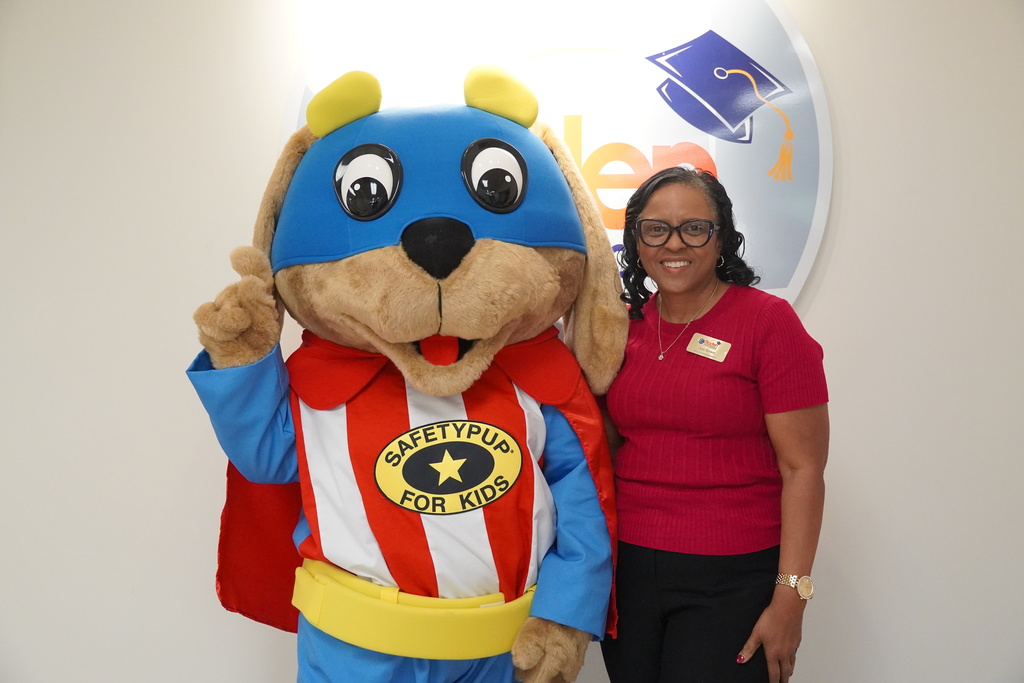 A staff member poses next to the Safety Pup mascot in front of the Bladen County Schools logo on the wall.