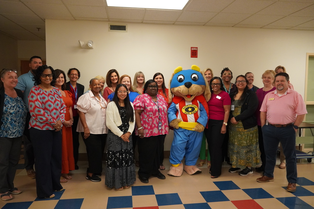 A large group of adults from Bladen County Schools stands together indoors, smiling with the Safety Pup mascot in the center wearing a red-and-white striped shirt, blue pants, and a cape.