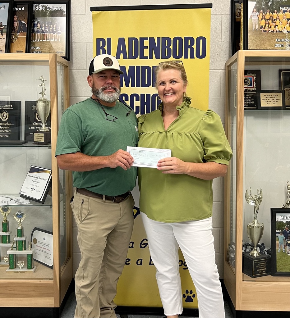 man and woman in green tops in front of a Bladenboro Middle Bulldogs sign holding a check and smiling