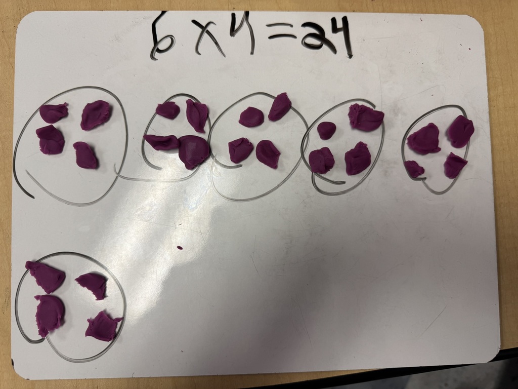 Close-up of a whiteboard showing purple Play-Doh pieces in groups inside circles, labeled with the multiplication equation 6 x 4 = 24.