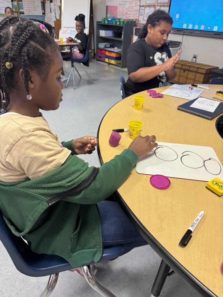 A student with braided hair uses a whiteboard and Play-Doh to create arrays for multiplication problems.