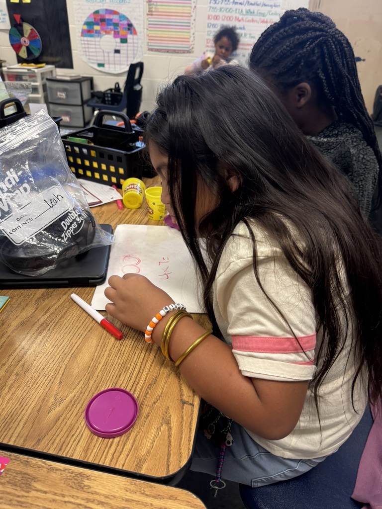 A student writes multiplication problems on a whiteboard while shaping purple Play-Doh pieces to represent an array.