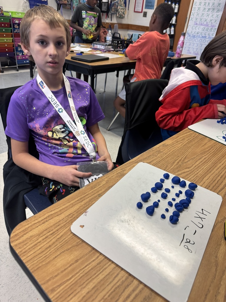 A student in a purple shirt shows a completed array with rows of blue Play-Doh pieces on a whiteboard labeled 4 x 7 = 28.