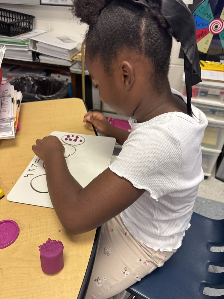 A student with a large black bow arranges purple Play-Doh pieces into circles drawn on a whiteboard.