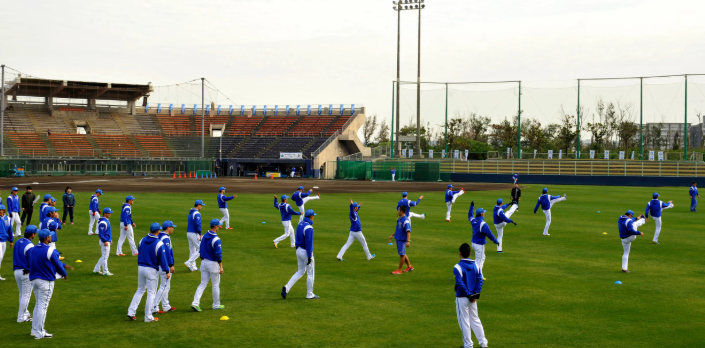 A large group of baseball players in blue jackets and white pants are spread across a baseball field doing warm-up exercises. Some players are stretching their legs while others are standing and watching. The background shows stadium seating, tall light poles, and a netted fence surrounding the field.