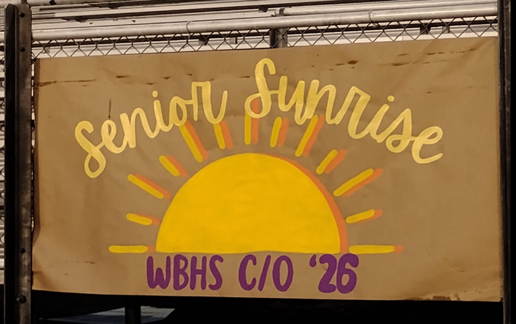 A handmade banner with a large yellow sun and orange rays painted on brown paper. Above the sun, in large yellow script, it says “Senior Sunrise.” Below the sun, in bold purple letters, it reads “WBHS C/O ’26.” The banner is attached to a chain-link fence with bleachers visible in the background.