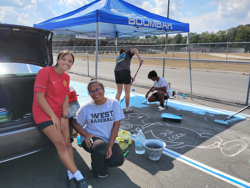Four students are painting and decorating a parking spot on asphalt. Two girls in the foreground smile at the camera; one is wearing a red shirt, and the other is wearing a gray “West Baseball” shirt. Behind them, one student uses a paint roller to spread blue paint while another kneels to outline a cartoon character labeled “Joe Cool.” A blue canopy tent provides shade, and the school stadium bleachers are visible in the background.