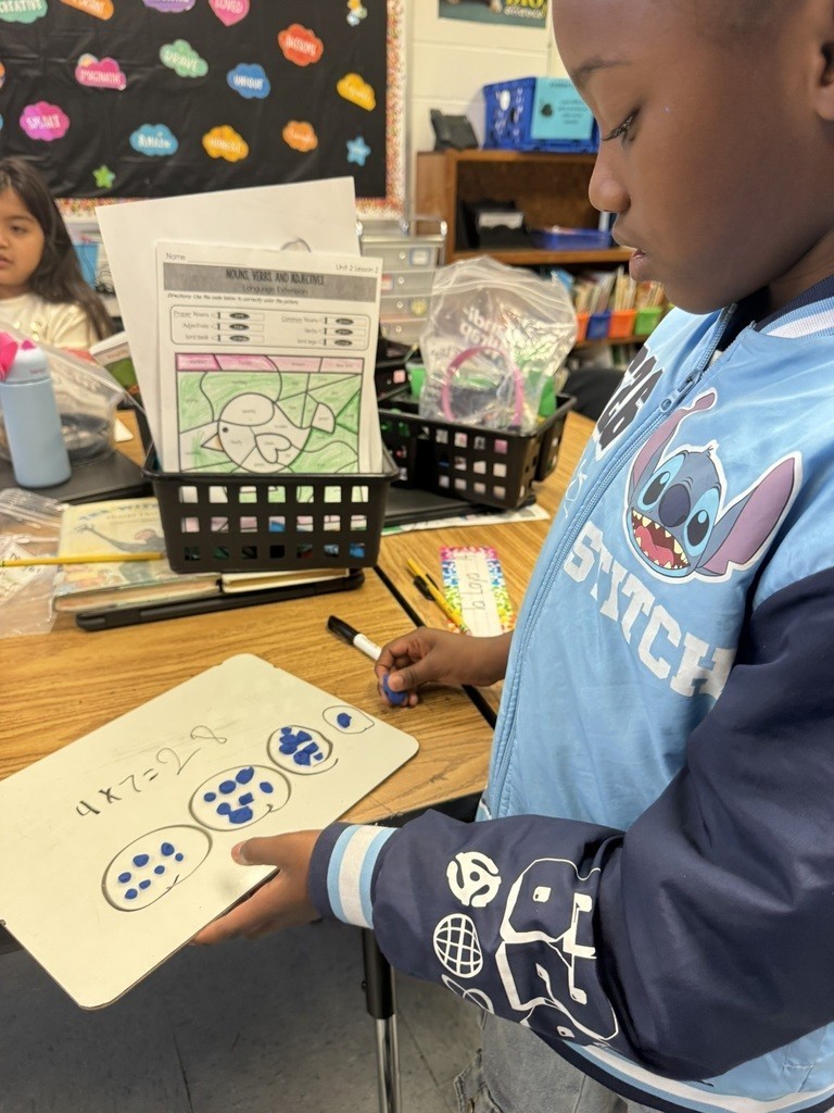 A student in a blue Stitch jacket holds a whiteboard showing a multiplication problem with groups of dots drawn.