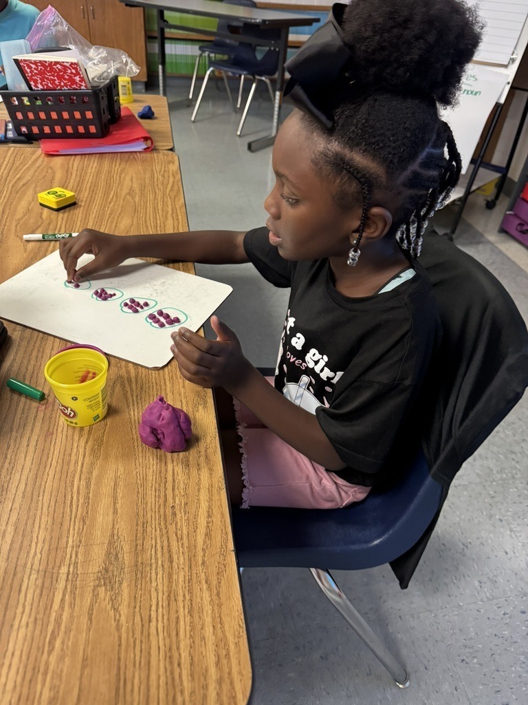 A student with a black bow in her hair arranges small Play-Doh pieces on a whiteboard to solve a multiplication problem.