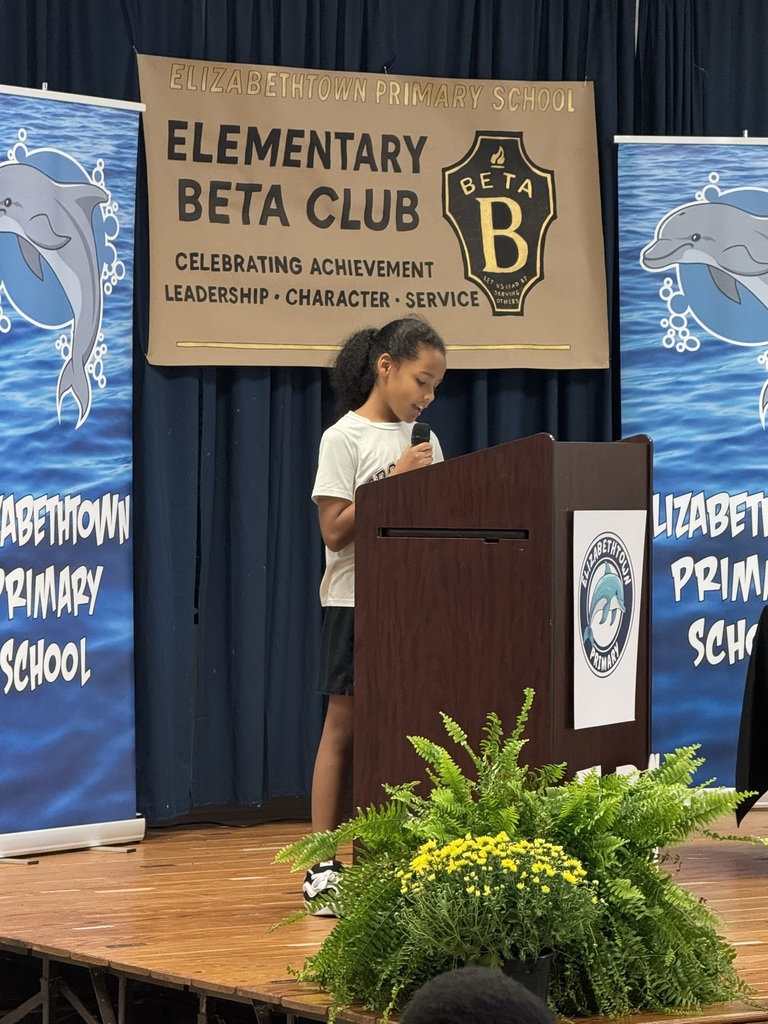 A student stands at a podium during an Elementary Beta Club ceremony, with banners and a decorative plant in the background.