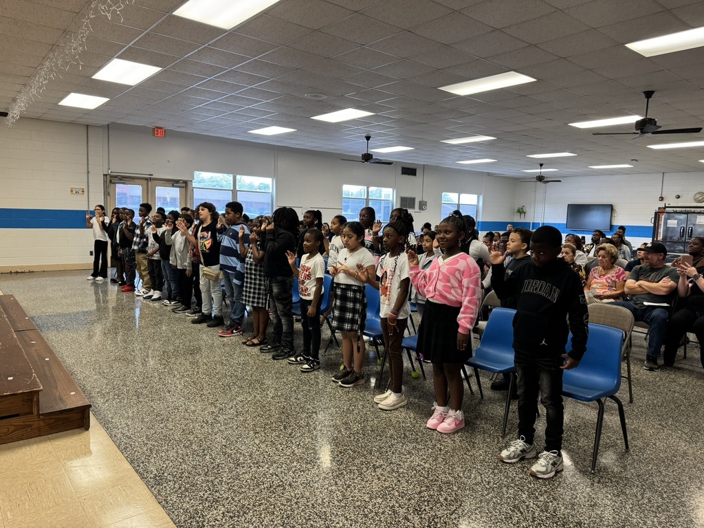 A group of children stand in a line, facing a stage in a school auditorium, participating in an event while adults watch from chairs.